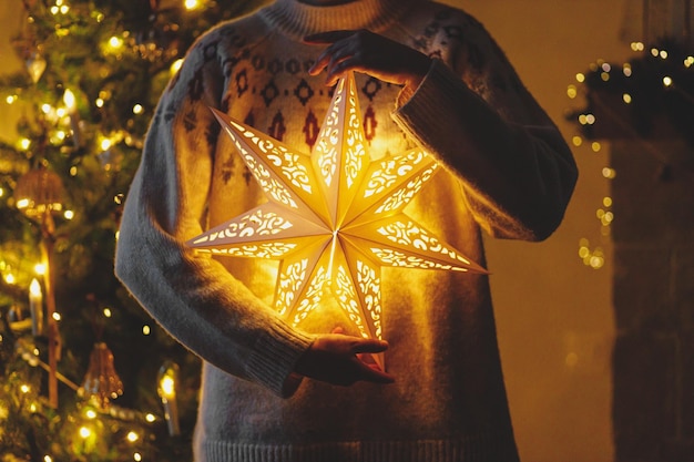 Mains dans un pull confortable tenant une étoile de Noël illuminée dorée sur fond d'arbre de Noël décoré avec style avec des lumières dorées dans la salle du soir Veille magique atmosphérique Joyeux Noël