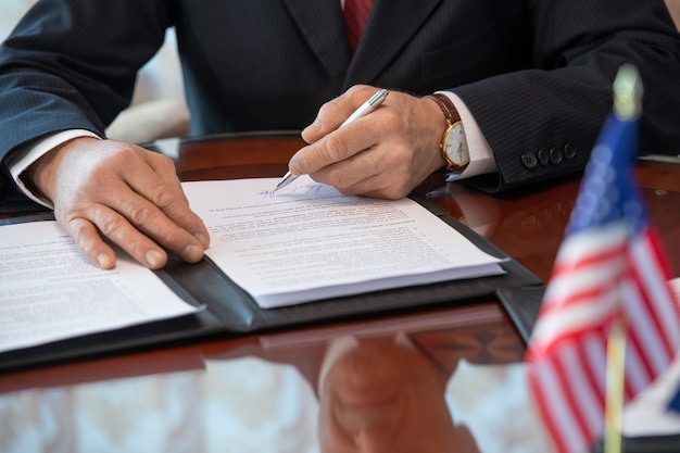 Mains d'un délégué américain mature en tenue de soirée signant un contrat de partenariat commercial et pointant vers la signature alors qu'il était assis près d'une table