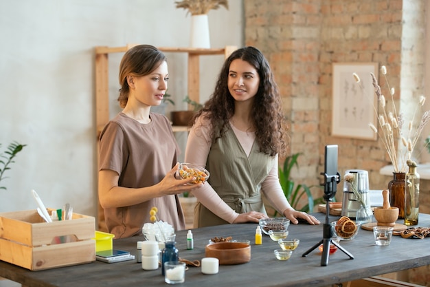 Mains de deux jeunes femmes coupant la masse de savon dur en cubes sur des planches en se tenant debout près d'une table avec des ingrédients pour la fabrication de produits cosmétiques