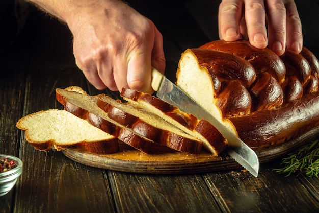 Les mains du chef coupées avec un couteau pain frais ou kalach sur une planche à découper de cuisine pour le petit déjeuner nourriture saine et concept de boulangerie traditionnelle