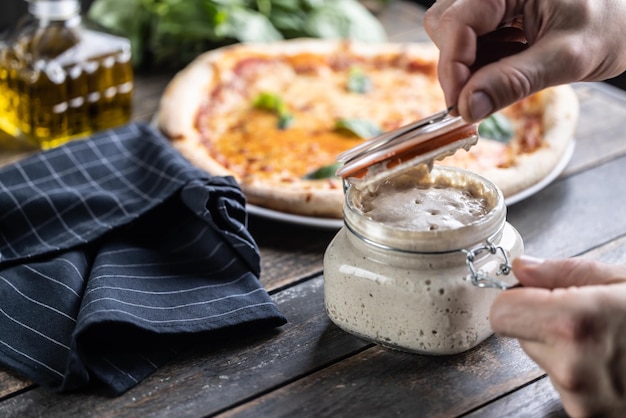 Les mains du cuisinier ouvrent un pot de levain de seigle en arrière-plan une pizza au levain