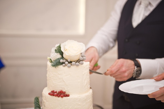 Les mains du marié coupent un gâteau de mariage blanc avec des fleurs et des baies
