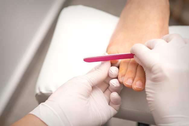 Les mains du pédicure dans des gants de protection en caoutchouc déposent les ongles sur les pieds avec une lime à ongles dans un salon de beauté.