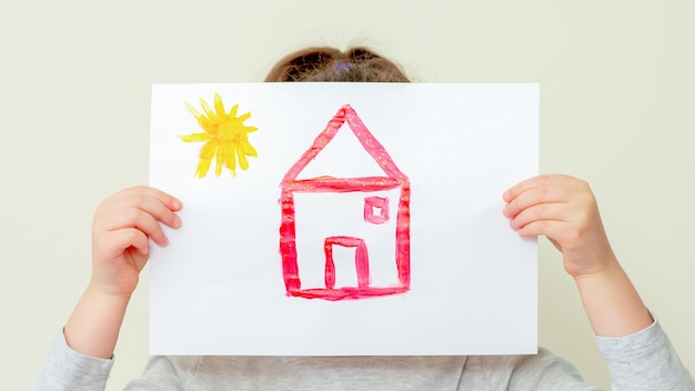 Mains d'enfant tenant une photo de maison couvrant son visage sur fond clair.