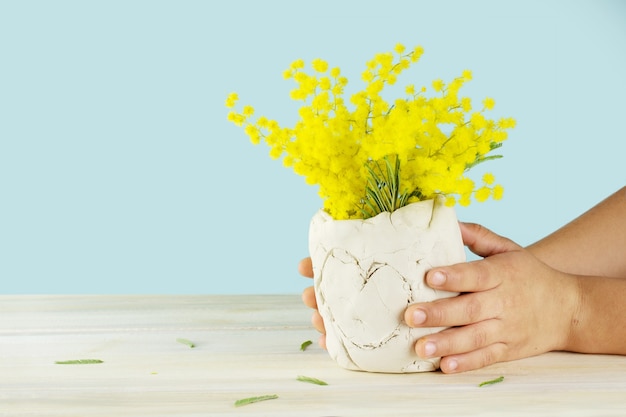 Les mains de l'enfant tenant un vase en céramique bricolage avec des fleurs jaunes. Isolé sur une surface bleue.