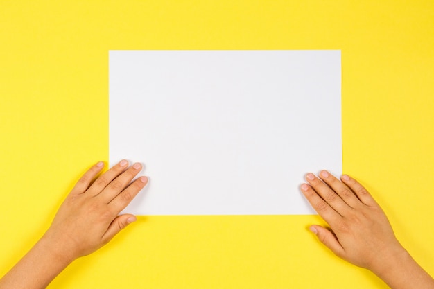 Mains d'enfants avec une feuille de papier vierge blanche sur fond jaune