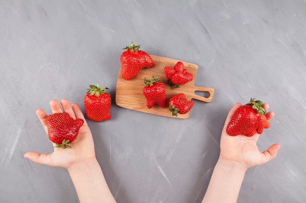 Les mains des enfants tiennent des fraises biologiques aux formes amusantes. Planche de service en bois avec des baies laides lumineuses.