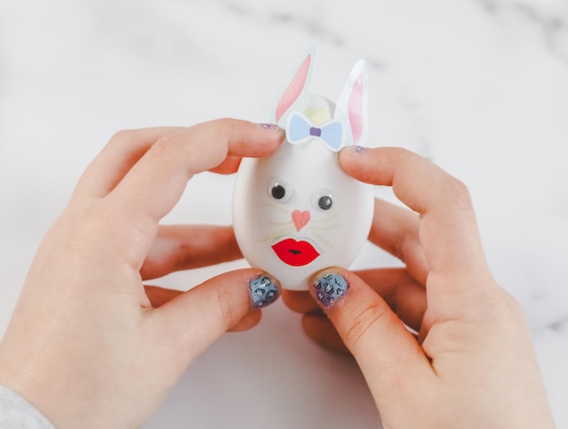 Les mains des enfants tiennent un œuf blanc avec des yeux collés, des lèvres, un nœud papillon et des oreilles de lapin en papier collé