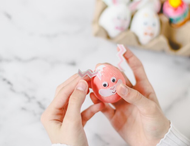 Les mains des enfants tiennent un œuf rose avec des yeux collés et collent des autocollants avec des oreilles de lapin en zigzag