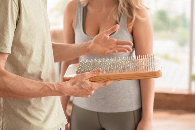 Mains d'un entraîneur de fitness masculin contemporain tenant un coussin de yoga avec des ongles tout en consultant l'un de ses clients dans un centre de sport et de remise en forme