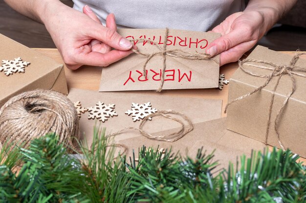 Photo des mains enveloppent une lettre de noël dans une enveloppe avec les mots joyeux nol sur la table avec des cadeaux de noël, de la ficelle et des flocons de neige. emballage pour les cadeaux de noël et du nouvel an.