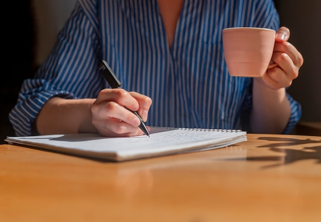 Mains d'étudiant écrivant avec un stylo dans le bloc-notes prenant des notes et tenant une tasse de café dans les mains