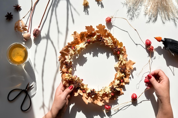 Mains faisant une couronne de fleurs séchées à partir de feuilles d'automne sèches et de baies d'automne. Les mains dans un pull avec des ongles manucurés fixent les décorations. Mise à plat sur une table blanche, lumière du soleil avec de longues ombres.