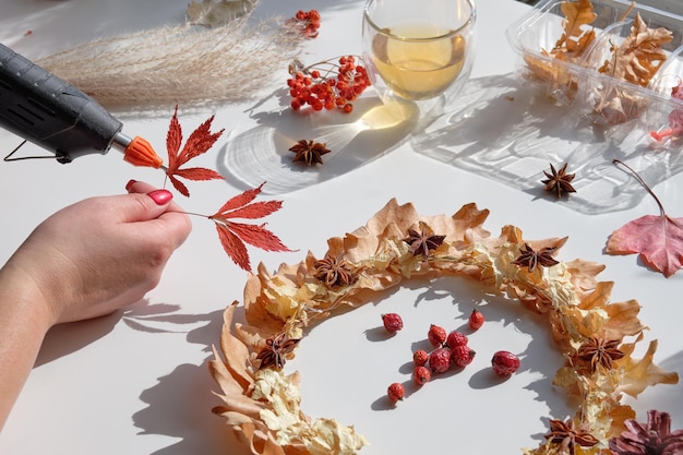 Mains faisant une couronne de fleurs séchées à partir de feuilles d'automne sèches et de baies d'automne Les mains avec des ongles manucurés fixent les décorations avec un pistolet à colle