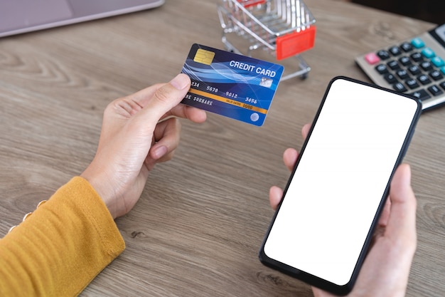 Mains féminines en chemise jaune tenant un téléphone portable avec un écran vide et une carte de crédit sur le bureau et utilisant un téléphone portable pour faire des achats en ligne, des opérations bancaires en ligne,