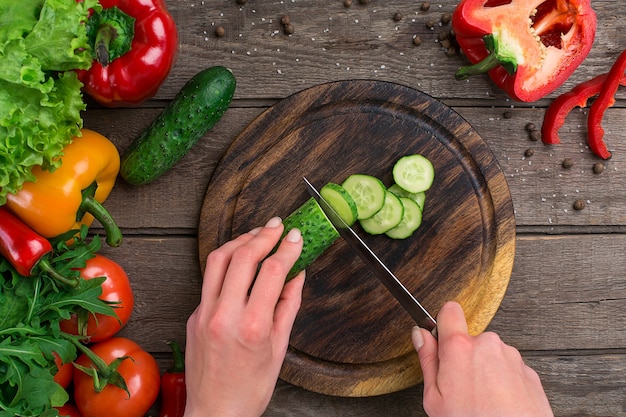 Mains féminines coupant le concombre à table, vue de dessus. Sur la table des légumes et une planche de bois