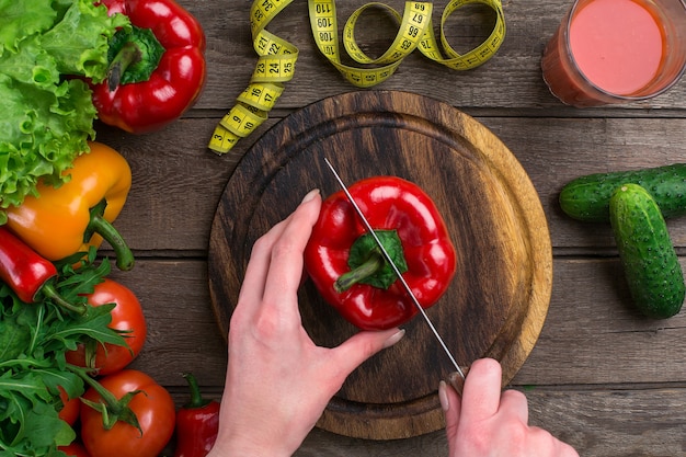 Mains féminines coupant le poivre à table, vue de dessus. Sur la table des feuilles de laitue, du poivre, un verre de jus de tomate, une planche de bois et un couteau