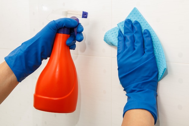 Des mains féminines dans des gants en caoutchouc bleu tiennent une bouteille de détergent et une serviette en microfibre. Une femme lave un mur avec des carreaux blancs dans la salle de bain. Outils et équipements de nettoyage