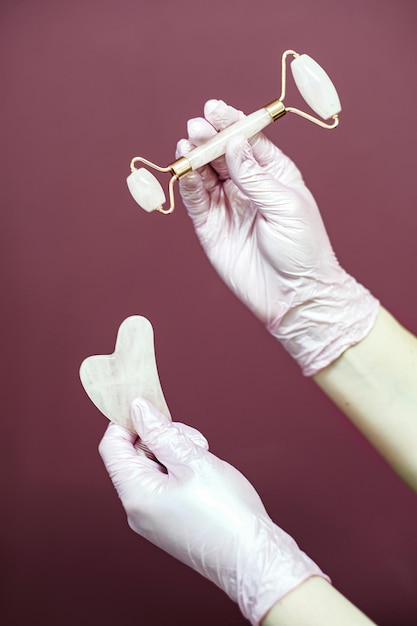 Photo mains féminines dans des gants roses tenant un rouleau et un grattoir gua sha sur fond sombre. copie espace