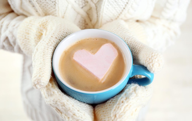 Mains féminines dans des mitaines chaudes tenant une tasse de cappuccino chaud avec guimauve coeur gros plan