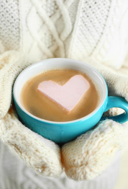 Mains féminines dans des mitaines chaudes tenant une tasse de cappuccino chaud avec guimauve coeur gros plan