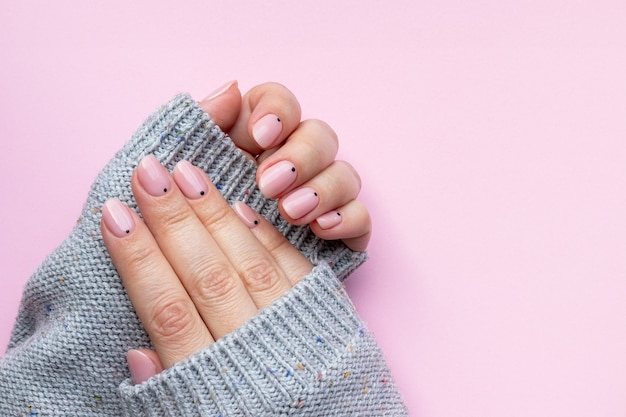 Mains féminines dans un pull en tricot gris avec une belle manucure à la mode - ongles nude roses avec de petits points noirs sur fond rose avec espace de copie