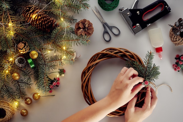 Des mains féminines décorent la couronne de Noël avec des branches d'épinette avec une baie rouge et des cônes forestiers. Fond blanc. Concept du nouvel an.