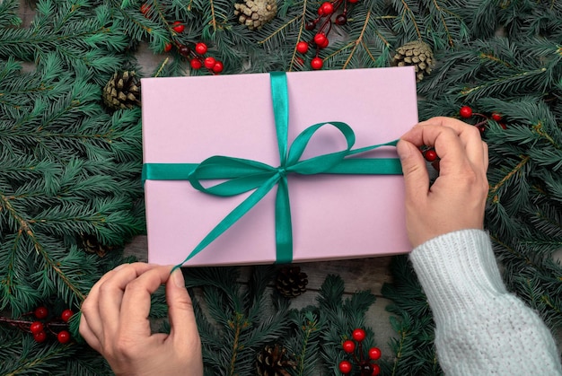 Les mains féminines détachent le ruban du cadeau de Noël sur fond de branches d'épinette sombre Cadeau de célébration de Noël enveloppé dans du papier rose sur fond de branches de sapin vue de dessus