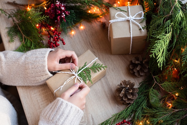 Mains féminines enveloppant un cadeau dans du papier kraft sur une table avec décor et guirlande de lumières, concept de Noël, gros plan.