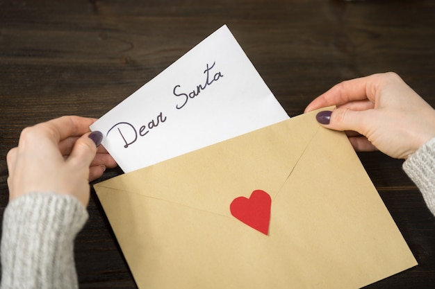Des mains féminines mettent une lettre pour le Père Noël dans une enveloppe. Fond en bois. Fermer