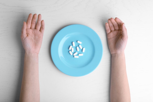 Des mains féminines et un plat rempli de pilules sur une table blanche.