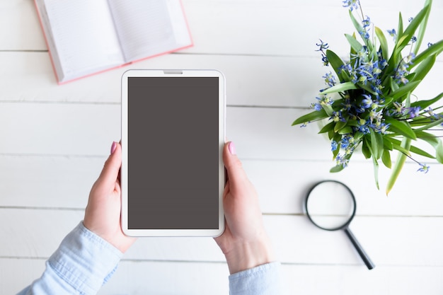 Mains féminines avec une tablette. Écran vide noir. Table avec carnet et fleurs