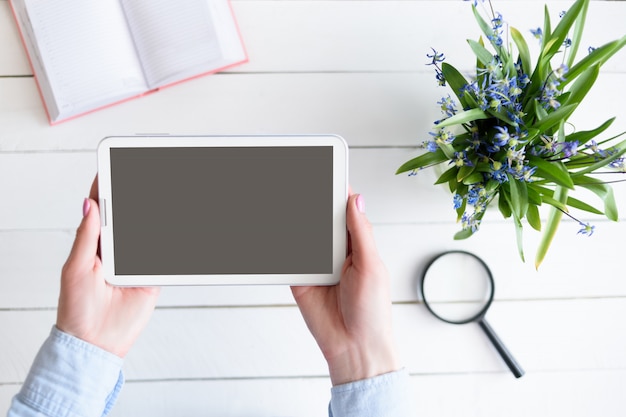 Mains féminines avec une tablette. Écran vide noir. Table avec carnet et fleurs