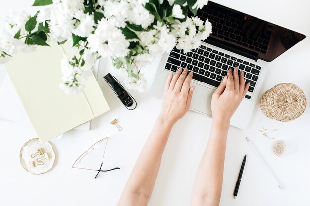 Mains féminines tapant sur ordinateur portable. Espace de travail de bureau avec ordinateur, bouquet de fleurs et papeterie sur tableau blanc