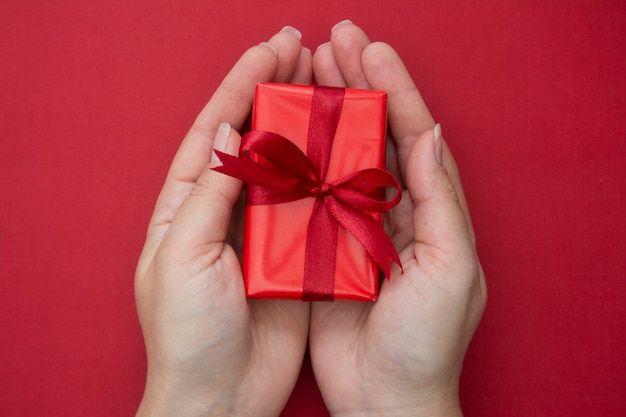 Mains féminines tenant une boîte de cadeau de Noël rouge avec ruban rouge et un arc, sur fond rouge.