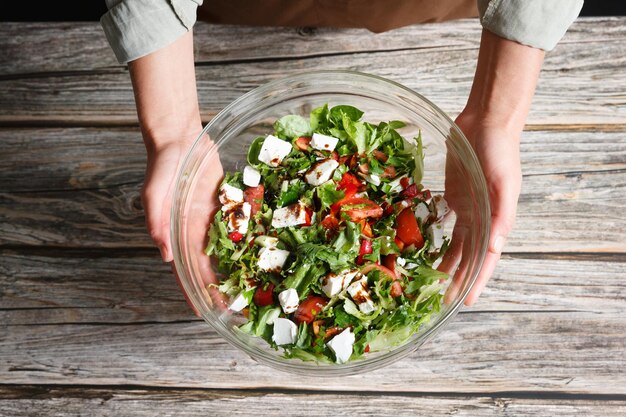 Des mains féminines tenant un bol en verre de salade de légumes avec de la feta sur un fond en bois