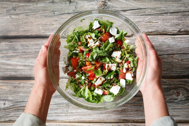 Des mains féminines tenant un bol en verre de salade de légumes avec de la feta sur un fond en bois