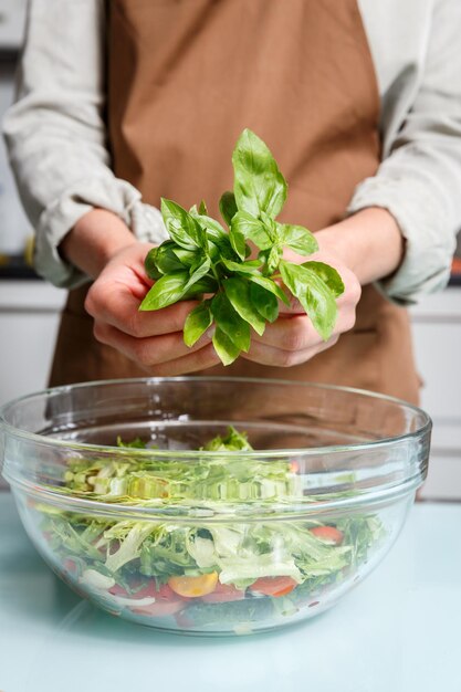 Des mains féminines tenant un bouquet de basilic vert sur un bol en verre avec de la salade sur le fond d'une cuisine