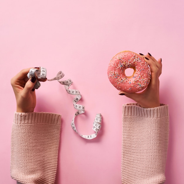 Mains féminines tenant donut et ruban à mesurer sur fond rose.