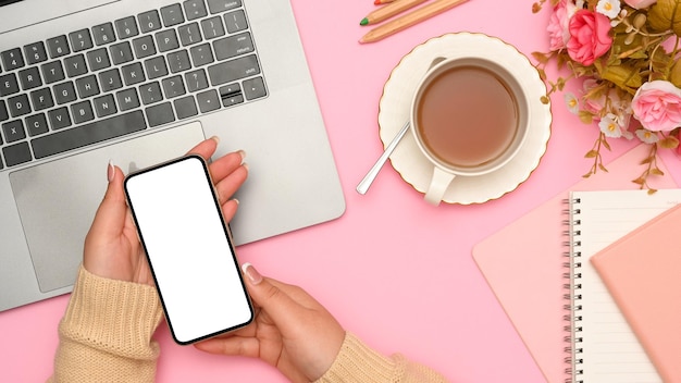 Mains féminines tenant une maquette d'écran blanc de smartphone sur sa vue de dessus de bureau rose élégant