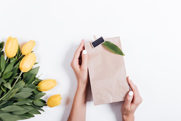 Mains féminines tenant un sac en papier avec un cadeau près du bouquet de Tulipes jaunes sur fond blanc