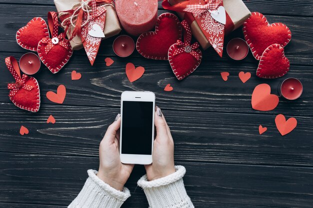 Mains féminines tenant le smartphone sur un fond avec des cadeaux, coeurs décoratifs pour la Saint-Valentin.