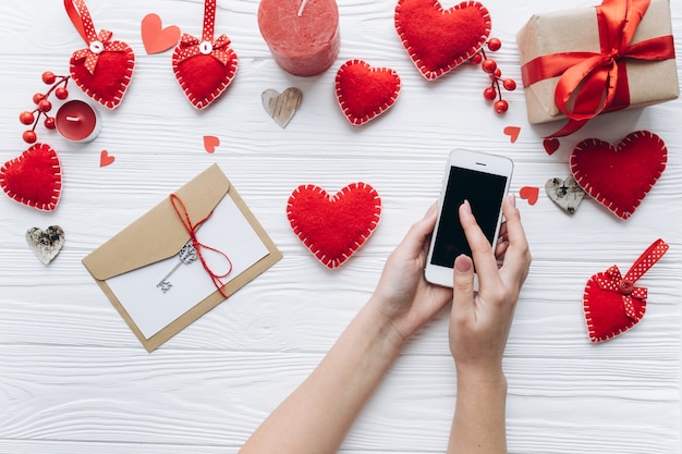 Mains féminines tenant le smartphone sur une table blanche avec des coeurs décoratifs pour la Saint-Valentin.