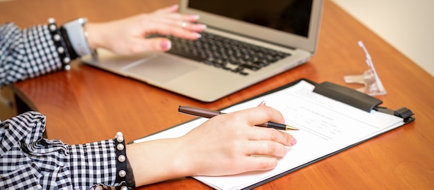 Mains féminines tenant un stylo sous le document et travaillant avec un ordinateur portable à la table dans un bureau