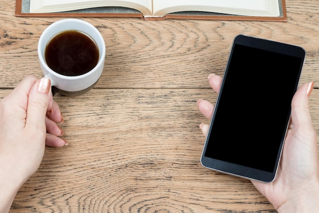 Mains féminines tenant une tasse de café et un smartphone noir sur le fond d'une table en bois avec un livre posé dessus