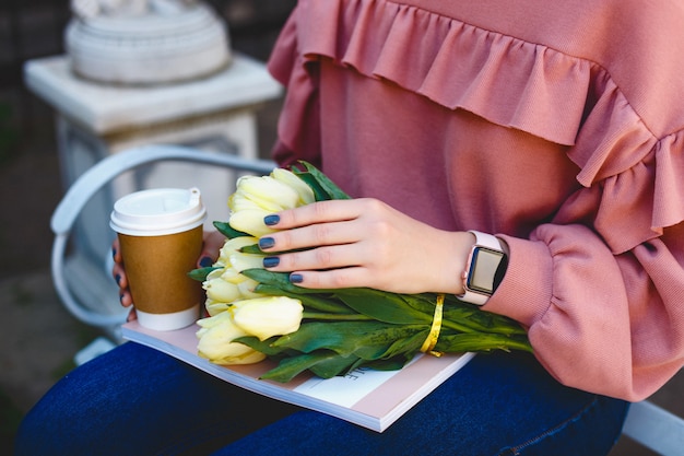 Mains féminines tenant un verre avec du café, des tulipes jaunes et un magazine