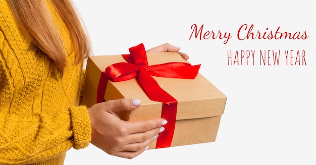 Des mains féminines tiennent une boîte-cadeau artisanale avec un arc rouge Noël, nouvel an. Espace de copie isolé. Une femme dans un pull jaune chaud confortable sans visage. Texte. Fond clair