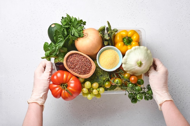 Des mains féminines tiennent une boîte en plastique avec un assortiment de légumes frais, de fruits, de céréales et de graines sur un fond en bois ancien. Livraison à domicile en toute sécurité, cuisson des aliments et arrière-plan et maquette d'aliments sains et propres.