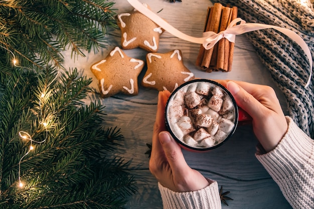 Des mains féminines tiennent une tasse de café ou de thé au-dessus d'une guimauve. Composition de Noël ou du nouvel an de boisson chaude, biscuits au gingembre et cannelle. Vue de dessus.