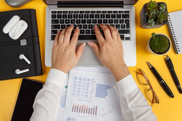 Mains de femme d'affaires tapant sur un ordinateur portable et travaillant avec des rapports vue de dessus sur un bureau jaune